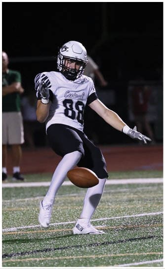 A person in a white football uniform is kicking a football on a field at night with several people standing in the background. Descripci n generada con IA