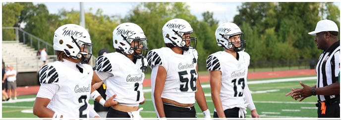 Four people in white football uniforms with helmets stand on a field listening to a person in a referee uniform. Descripci n generada con IA