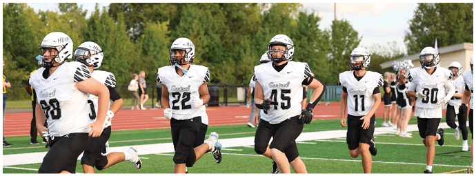 People wearing white football uniforms with helmets are standing on a track field with trees and cheerleaders in the background. Descripci n generada con IA