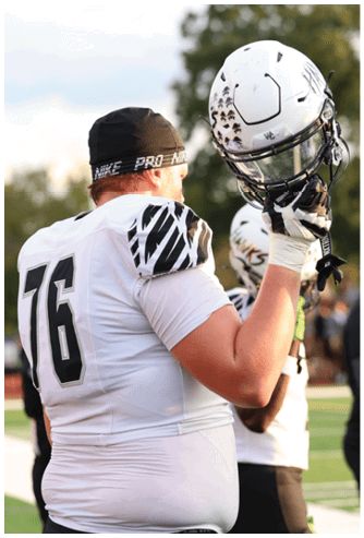 A person wearing a white sports uniform and green gloves is holding up a white football helmet with “HAWKS" written on it while other players and a coach stand in the background on a field. Descripci n generada con IA