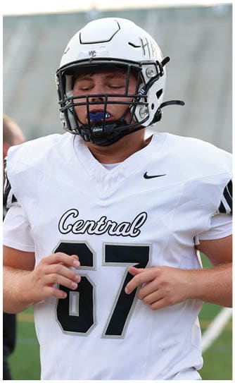 A person wearing a white football jersey and helmet is in the foreground while another person in a black vest and white shirt stands blurred in the background on a sports field. Descripci n generada con IA