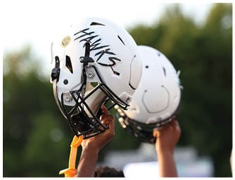 Hands are holding up white football helmets with the word “HAWKS" written on one of them against a blurred outdoor background. Descripci n generada con IA