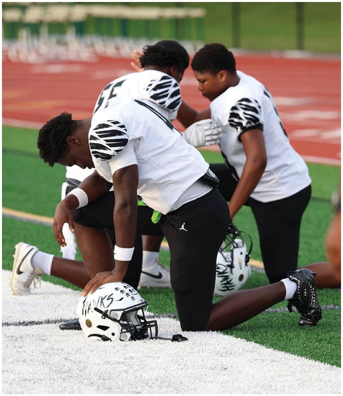Three people in football uniforms kneel on a field with their helmets on the ground, appearing to be in a moment of reflection or prayer. Descripci n generada con IA