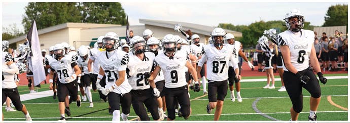 A group of people wearing white and black football uniforms are running on a green field with a crowd and cheerleaders in the background. Descripci n generada con IA