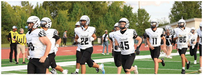 People wearing white football uniforms and helmets are walking on a track near a field with trees and a few other people in the background. Descripci n generada con IA