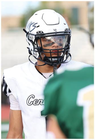 Two people wearing football helmets and uniforms are facing each other on a field, with one in a white jersey and the other in a green jersey. Descripci n generada con IA