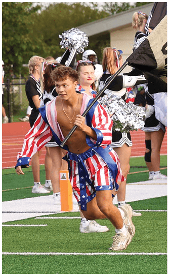 A group of people in football uniforms are running onto a field with cheerleaders in the background holding pom-poms. Descripci n generada con IA