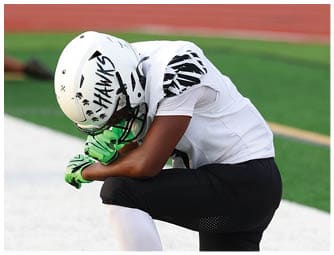 A person in football gear kneels on one knee on a field with others kneeling in the background near the sideline. Descripci n generada con IA