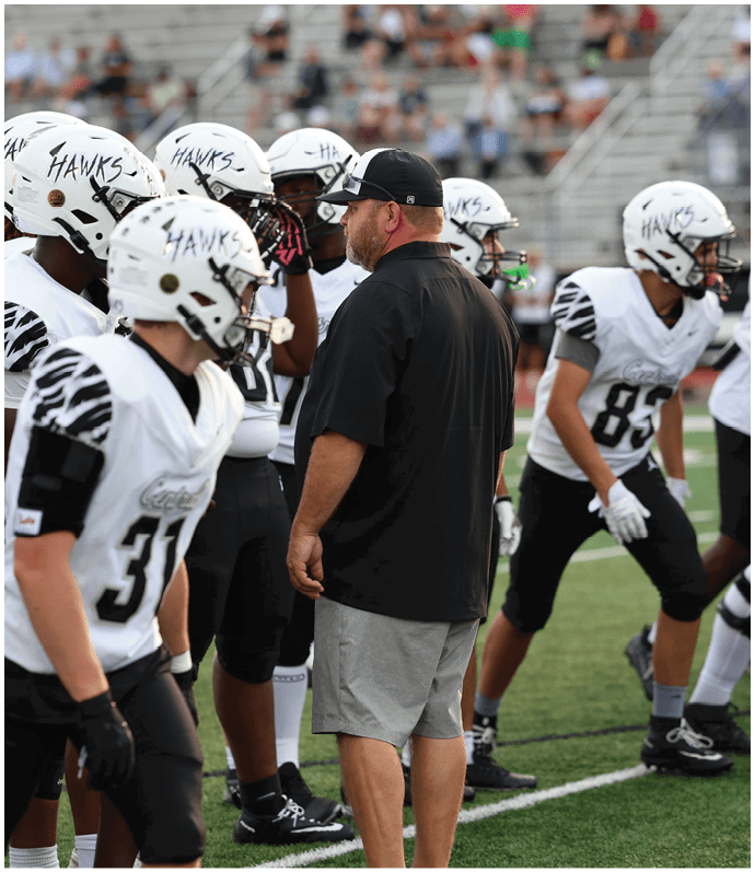 A group of people wearing white football helmets and uniforms with “HAWKS" written on the helmets are gathered on a sports field with one person in a black shirt and gray shorts standing among them. Descripci n generada con IA