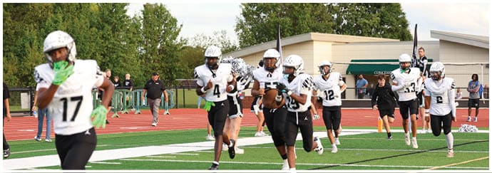 People in football uniforms and helmets are gathered on a field with trees and a building in the background. Descripci n generada con IA