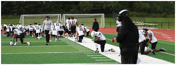 People in sports uniforms are kneeling and stretching on a football field near the sidelines while a person in dark clothing stands nearby. Descripci n generada con IA