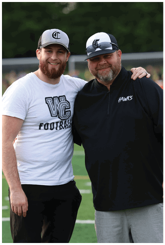 Two adults wearing baseball caps smile with their arms around each other on a sports field, one wearing a white shirt with “WC Football" and the other in a black jacket. Descripci n generada con IA