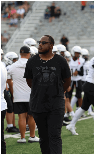 A person wearing sunglasses and a black t-shirt with a skull design stands on a sports field with players in white helmets and uniforms in the background. Descripci n generada con IA