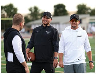 Three adults wearing sportswear and caps are standing on a football field, with one holding a football and all appearing engaged in conversation. Descripci n generada con IA