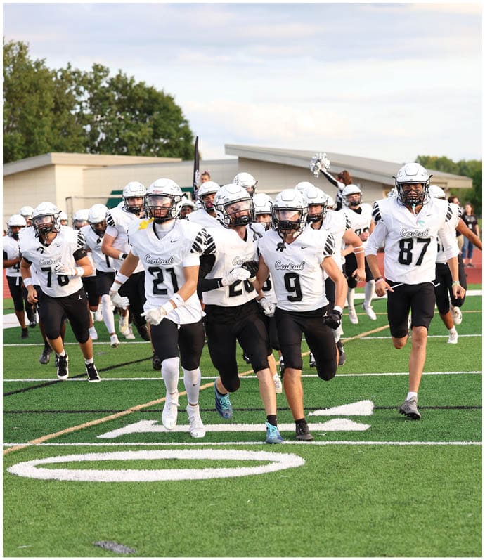 People in football uniforms are running on a green field with a crowd and cheerleaders in the background. Descripci n generada con IA