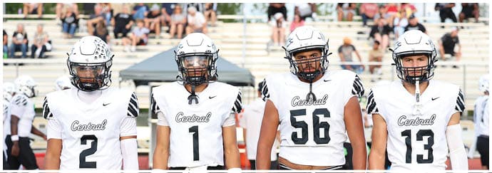 Four people wearing white football uniforms with helmets stand side by side on a football field with spectators in the background. Descripci n generada con IA