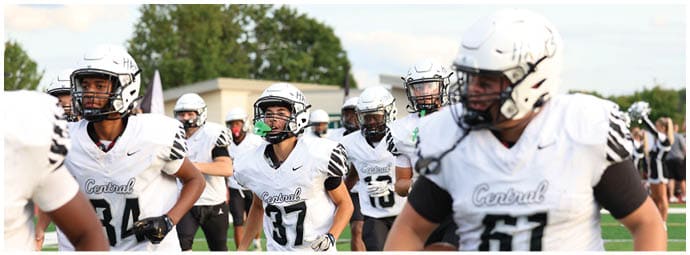 People wearing white and black football uniforms are running on a field, with one person holding a football. Descripci n generada con IA