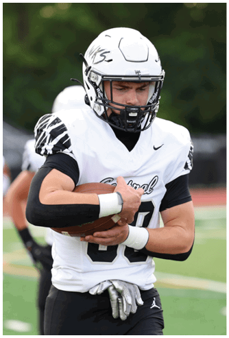 A person wearing a white football helmet and uniform is holding a football close to their chest on a sports field. Descripci n generada con IA