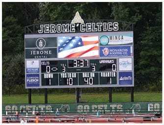 A football field with a scoreboard displaying the name Jerome Celtics and showing game information against a backdrop of trees and an American flag at half-mast. Descripci n generada con IA
