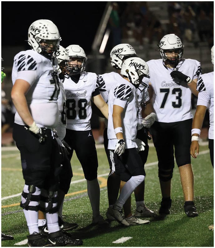 Several people in football uniforms and helmets stand on a field at night with a blurred group of people in the background. Descripci n generada con IA