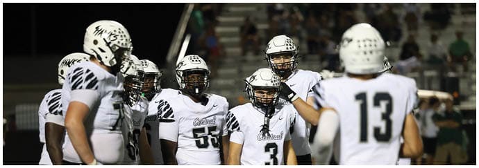 A group of people wearing white football uniforms with helmets are gathered on a field, with one person facing away from the camera. Descripci n generada con IA