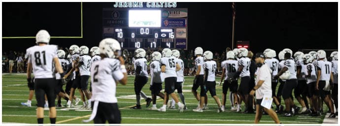 People in football uniforms are gathered on a field at night with a scoreboard in the background showing the game has ended. Descripci n generada con IA