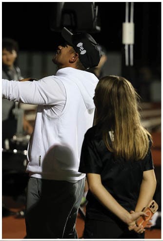 A person wearing a white sleeveless hoodie and a black cap is reaching out while another person with long hair and a black shirt stands nearby. Descripci n generada con IA