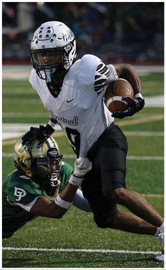 A person in a white football uniform with a helmet is running with a football while another person in a green football uniform attempts to tackle them on a field. Descripci n generada con IA