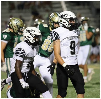 Two people in white football uniforms with “Hawks" helmets stand in the foreground while three people in green and gold football uniforms stand in the background on a field. Descripci n generada con IA