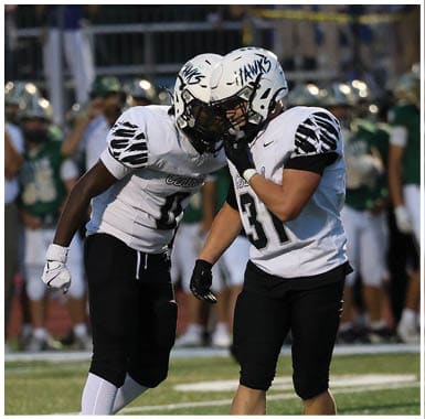 Two people in white and black football uniforms are standing close together on a field while a referee and other players in green uniforms are in the background. Descripci n generada con IA