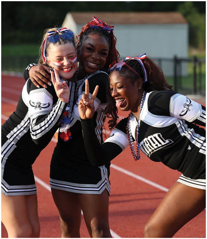 Three adults in cheerleading uniforms are smiling and posing with peace signs on an outdoor track. Descripci n generada con IA