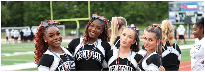 Four people wearing matching cheerleading uniforms and red, white, and blue accessories smile and pose together outdoors on a sports field. Descripci n generada con IA