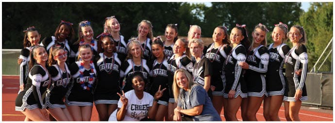 A group of people wearing matching cheerleading uniforms pose together on a track field with trees in the background. Descripci n generada con IA