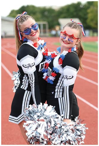 Two people in matching black and white cheerleading uniforms with colorful leis and heart-shaped sunglasses smile while holding silver pom-poms on a running track. Descripci n generada con IA