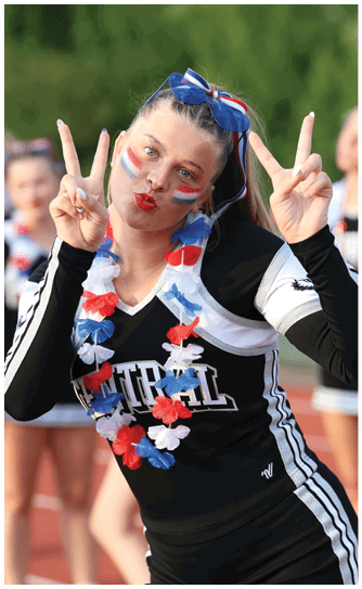 A person in a black and white cheerleading uniform with red, white, and blue face paint and a matching flower lei is making peace signs with both hands while standing on a track with other cheerleaders in the background. Descripci n generada con IA