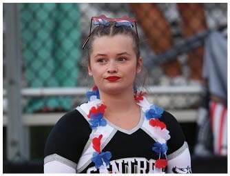 A person wearing a black and white cheerleading uniform with the word “CENTRAL" and a red, white, and blue flower lei stands in front of a blurred background with a fence and people. Descripci n generada con IA