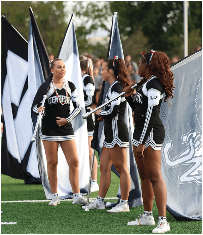 People in cheerleading uniforms stand on a football field holding large flags with team logos while others are visible in the background. Descripci n generada con IA