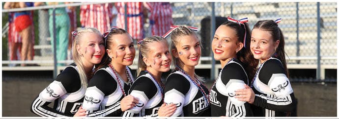 Six people wearing matching cheerleading uniforms with bows in their hair smile while standing close together outdoors. Descripci n generada con IA