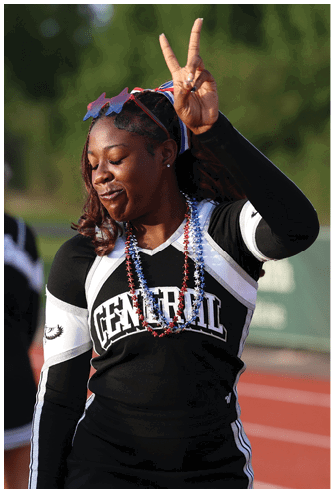 A person wearing a cheerleading uniform with “CENTRAL" on the front is making a peace sign with one hand and wearing star-shaped sunglasses and red and blue bead necklaces. Descripci n generada con IA