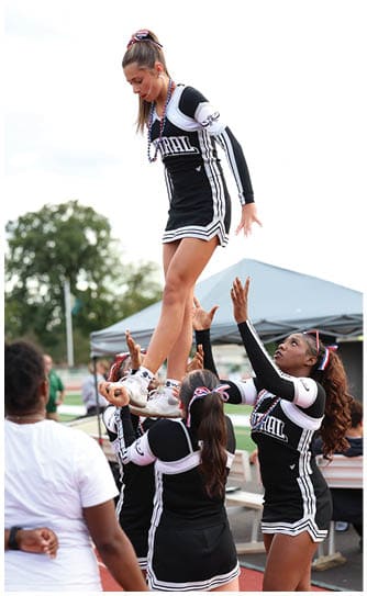 A person wearing a cheerleading uniform with a bow in their hair and beaded necklaces is looking downward outdoors. Descripci n generada con IA