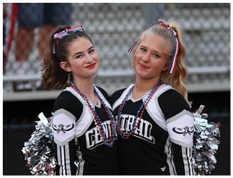 Two adults wearing matching cheerleading uniforms with “CENTRAL" on the front pose closely together with silver pom-poms and red, white, and blue hair ribbons and necklaces. Descripci n generada con IA