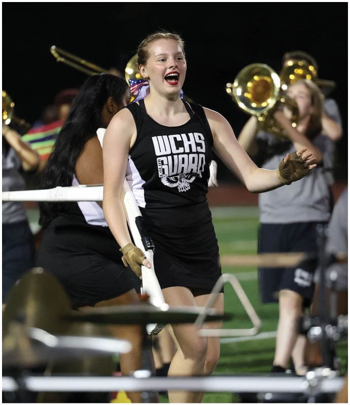 A person in a black and white uniform with “WCHS Guard" on the front is smiling and holding a white rifle while marching on a field with a band playing brass instruments in the background. Descripci n generada con IA