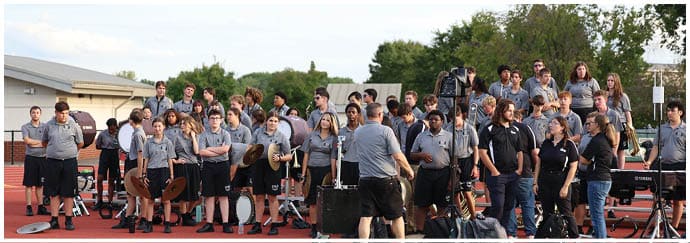A large group of people wearing matching gray and black shirts are gathered outdoors on a track field, some holding musical instruments and others standing or sitting on benches. Descripci n generada con IA