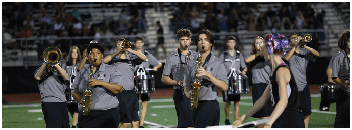 A group of people in matching gray shirts are playing brass and woodwind instruments while marching on a sports field with a person in a black outfit and colorful hair accessories leading them. Descripci n generada con IA