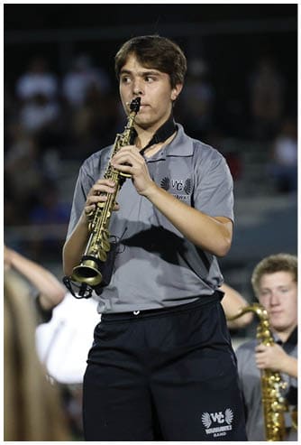 A person wearing a gray shirt and black shorts is playing a saxophone outdoors at night with another person in the background also holding a saxophone. Descripci n generada con IA