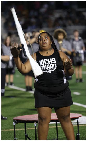 A person wearing a black WCHS Guard uniform is tossing a white rifle prop in the air on a sports field with other people blurred in the background. Descripci n generada con IA