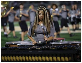 A person with long hair wearing a gray shirt is playing a marimba with mallets while a marching band performs in the background on a field. Descripci n generada con IA