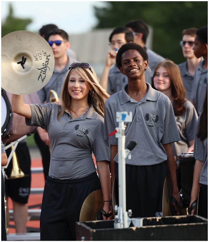 A group of people wearing matching gray shirts and black pants are playing musical instruments outdoors, with one person smiling and holding up a cymbal. Descripci n generada con IA