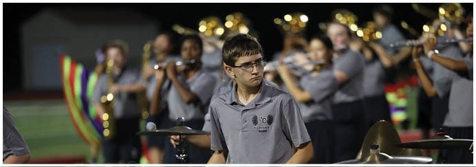A person wearing glasses and a gray polo shirt plays drums while a group of people in similar attire play flutes in the background on a field. Descripci n generada con IA