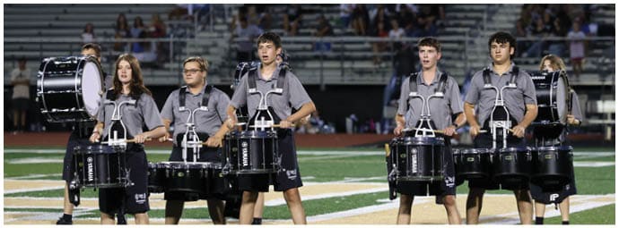 A group of people wearing matching gray shirts and black shorts are standing on a football field playing marching band drums with a crowd seated in the background. Descripci n generada con IA